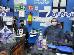 De izquierda a derecha Gerardo y [E2] en Ciber Plug. Se observan las señalizaciones con los que cuenta el negocio, el gel antobacterial y dos bandejas azules donde se sanitizan las monedas y los billetes. Fotografía: Ulises Chávez Valdovinos.