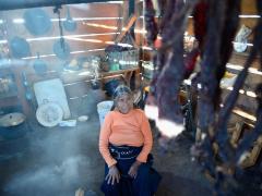 Mujer tzeltal sentada en cuarto de madera. Se observan algunos artículos del hogar y como entra la luz a través de los tabloides de madera. Fotografía: Jacobo García.