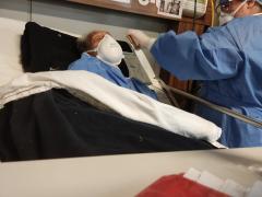 Paciente de la tercera edad con puntas nasales de alto flujo y cubrebocas kn95. La trabajora social le reproduce los mensajes de voz que los familiares grabaron para el paciente. En la pared se le observa una hoja de papel donde hay tres fotografías del paciente. Fotografía: Ulises Felipe Chávez Valdovinos