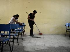 Mujer limpiando las bancas y hombre barriendo el piso de un salón de clases. por Aarón Álvarez.