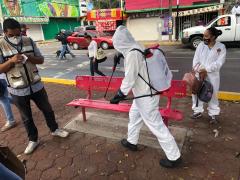 Brigadista sanitiza banca en un espacio público con personas al rededor. Fotografía: Facebook Territorial Santa Cruz-Quetzalcóatl Alcaldía Iztapalapa.